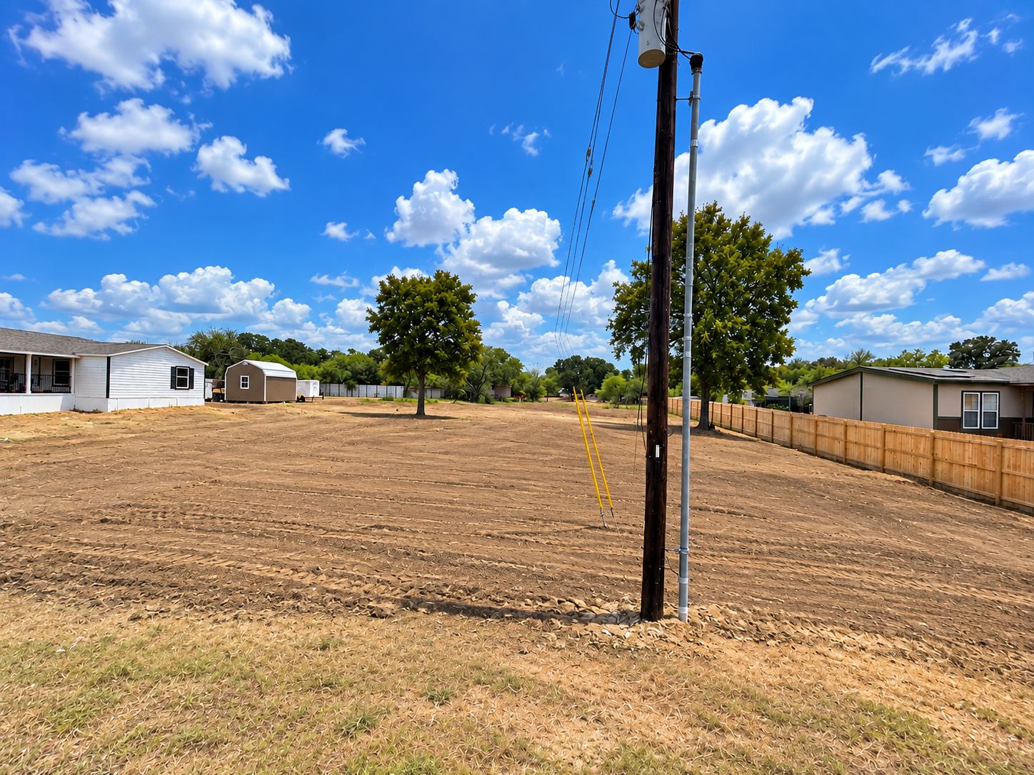 Cleared and graded lot with fresh tire tracks, a utility pole and homes in the background